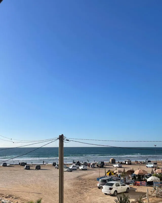 Anza beach seen from upstairs at Line Up: the beach car park, the sand, white water and the Atlantic horizon under a clear blue sky